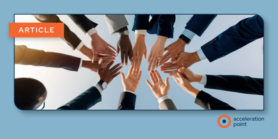 A group of professionals in business attire reaching their hands toward a central point from below, forming a circle of joined hands against a light background. The image represents cross-functional collaboration and team alignment -- core principles in