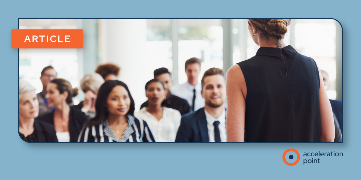 A presenter stands before an engaged audience in a professional conference setting, viewed from behind. Attendees in business attire face the speaker, suggesting a formal educational or scientific session. The image represents the in-person engagement and